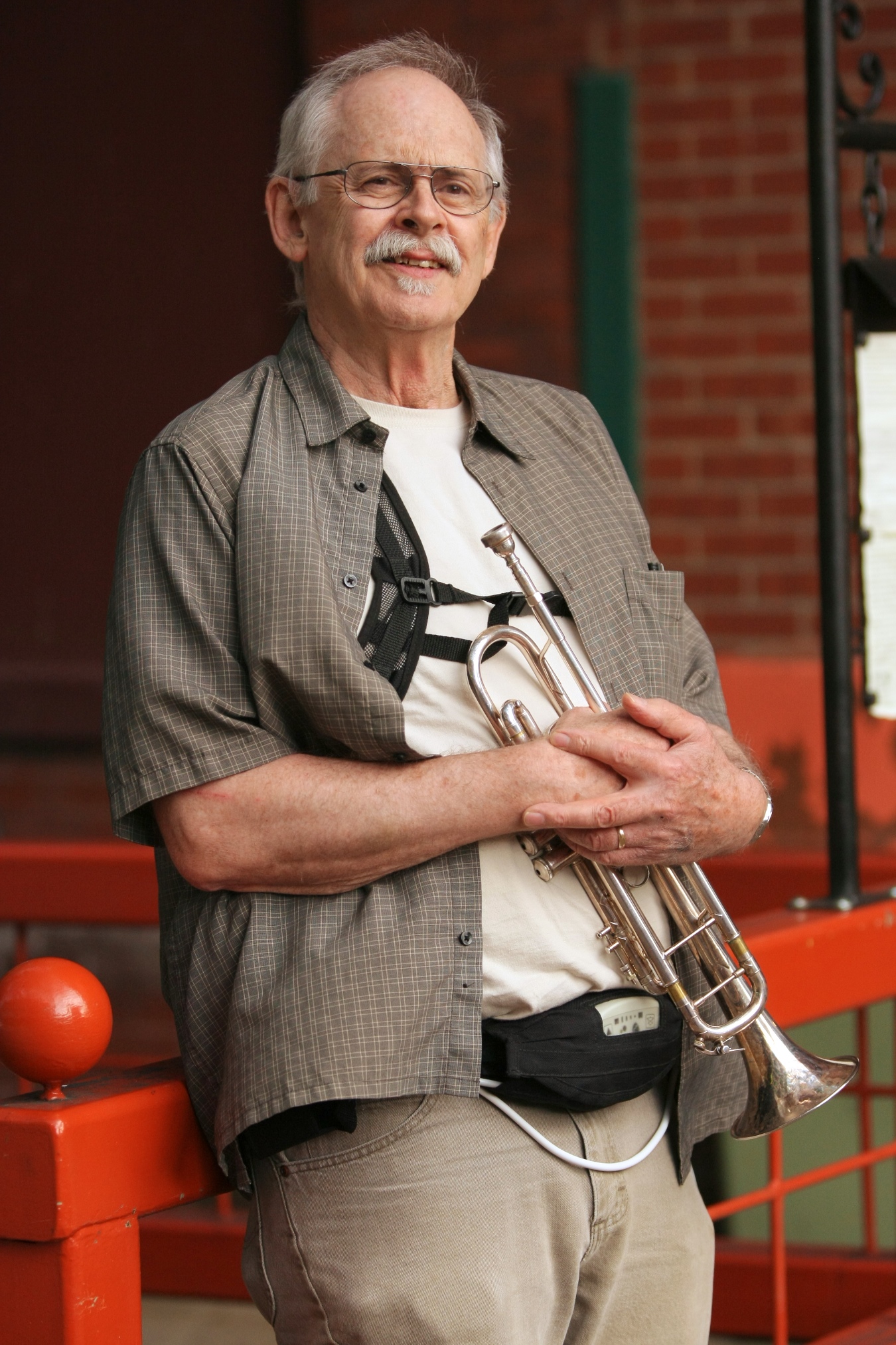 Paul Smoker, leaning up against a red metal fence, holding his trumpet; the external components of his LVAD are visible
